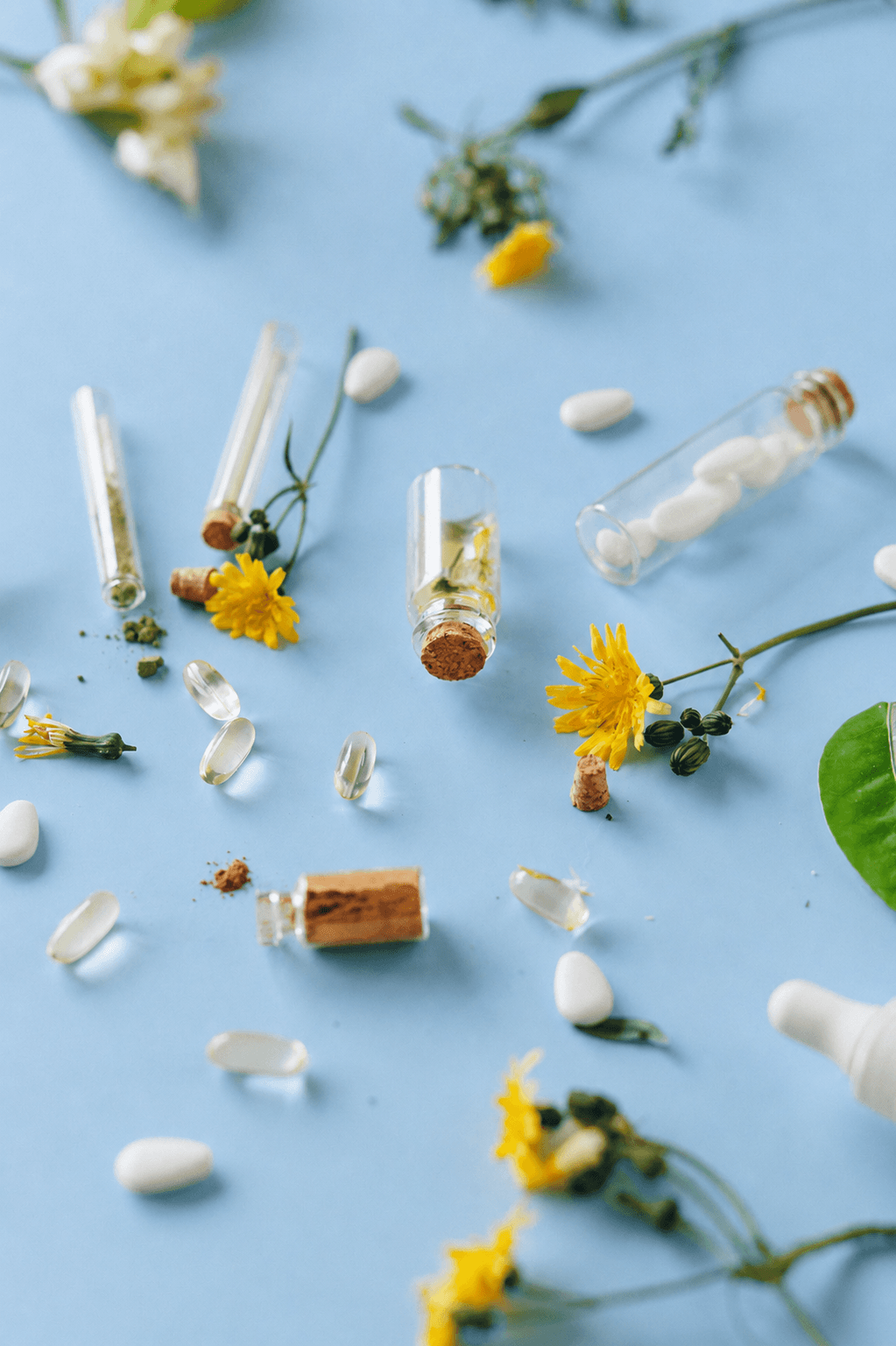 A flat lay of glass vials, capsules, powders, and yellow flowers on a light blue background